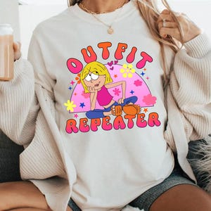 May include: A cream-colored crewneck sweatshirt featuring a graphic print of a cartoon character with blonde hair wearing a pink top and blue pants. The text OUTFIT REPEATER is printed in bold pink letters above and below the character illustration.