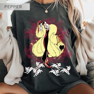 K&ouml;nnte beinhalten: Dunkelgraues T-Shirt mit einer Cartoon-Illustration von Cruella de Vil, die einen Dalmatiner-Welpen h&auml;lt, umgeben von laufenden Dalmatiner-Welpen. Das Design umfasst einen gelben Mantel und einen roten Hintergrund. Das Wort "PEPPER" befindet sich oben links.