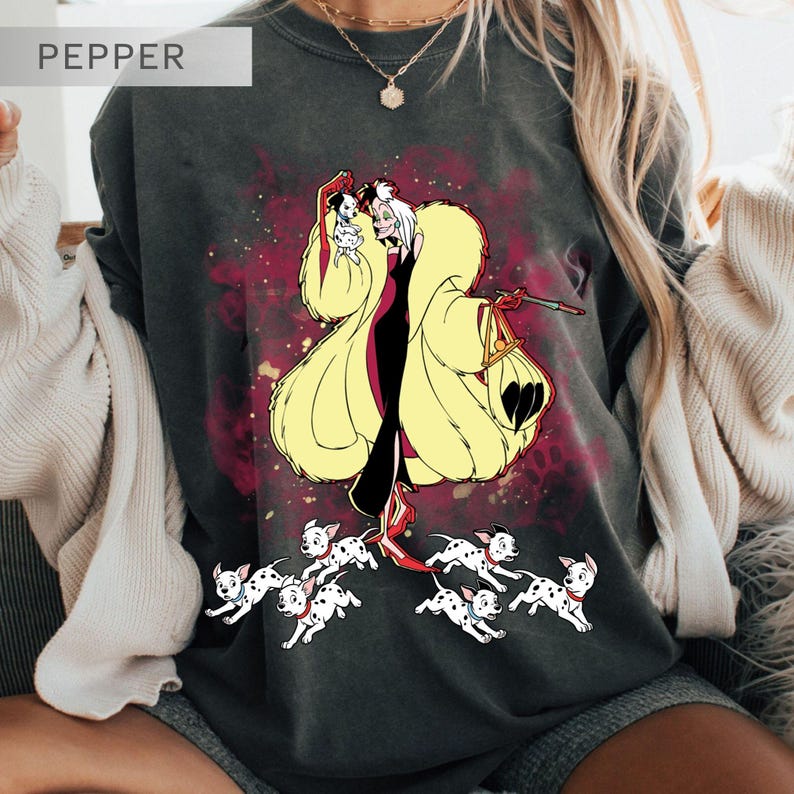 K&ouml;nnte beinhalten: Dunkelgraues T-Shirt mit einer Cartoon-Illustration von Cruella de Vil, die einen Dalmatiner-Welpen h&auml;lt, umgeben von laufenden Dalmatiner-Welpen. Das Design umfasst einen gelben Mantel und einen roten Hintergrund. Das Wort "PEPPER" befindet sich oben links.