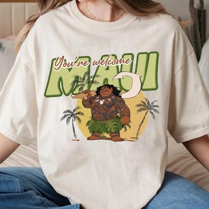 Puede incluir: Una camiseta blanca con un estampado gr&aacute;fico de Maui de la pel&iacute;cula de Disney Moana. El gr&aacute;fico presenta a Maui con su anzuelo, una palmera y el texto "You're welcome Maui".