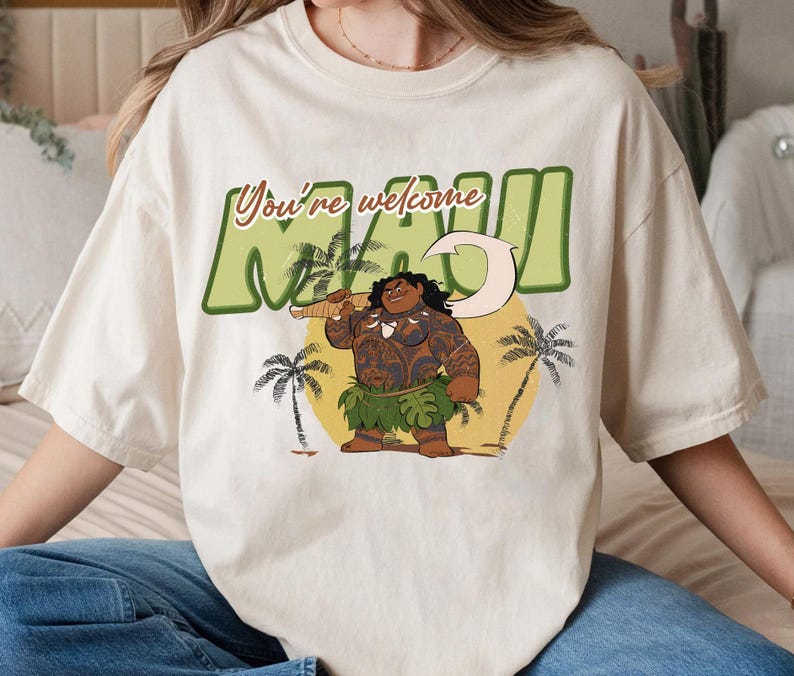 Puede incluir: Una camiseta blanca con un estampado gr&aacute;fico de Maui de la pel&iacute;cula de Disney Moana. El gr&aacute;fico presenta a Maui con su anzuelo, una palmera y el texto "You're welcome Maui".