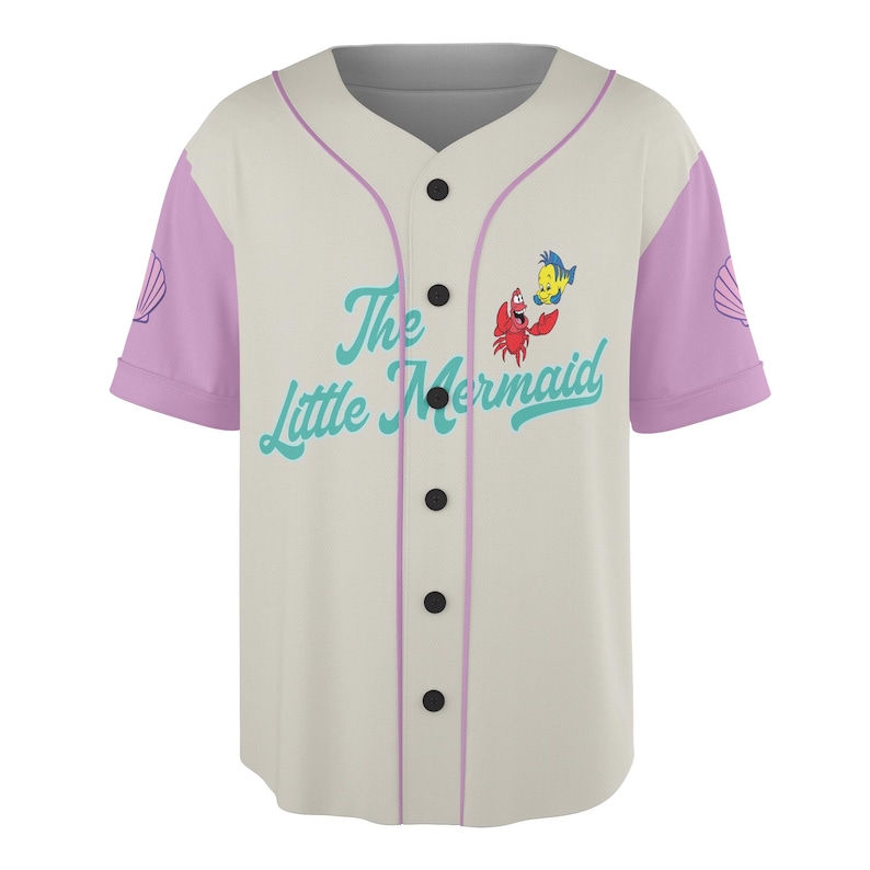 May include: Cream-colored baseball jersey featuring the text "The Little Mermaid" in teal script, along with cartoon images of Sebastian and Flounder. The sleeves are lavender with a seashell design. The jersey has black buttons and lavender trim.