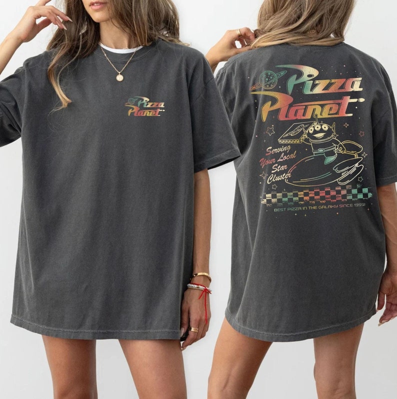 May include: An oversized dark grey t-shirt featuring a graphic print of the Pizza Planet logo and a character from the film Toy Story. The text on the shirt reads "Pizza Planet", "Serving Your Local Star Cluster", and "Best Pizza in the Galaxy Since 1995".