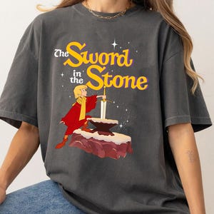 K&ouml;nnte beinhalten: Dunkelgraues T-Shirt mit einer Grafik aus "The Sword in the Stone". Das Design zeigt eine Figur in roter und gelber Kleidung, die ein Schwert aus einem Stein zieht. Der Text "The Sword in the Stone" ist gelb und wei&szlig;.