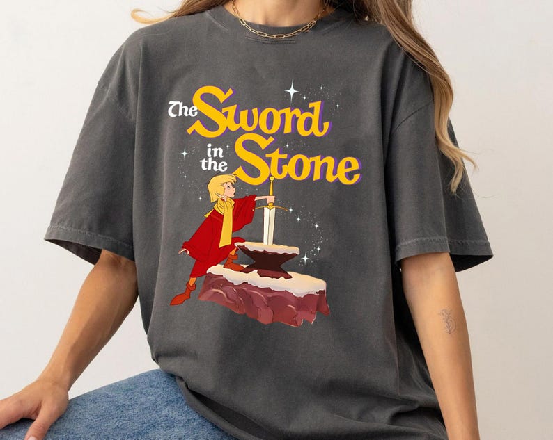 K&ouml;nnte beinhalten: Dunkelgraues T-Shirt mit einer Grafik aus "The Sword in the Stone". Das Design zeigt eine Figur in roter und gelber Kleidung, die ein Schwert aus einem Stein zieht. Der Text "The Sword in the Stone" ist gelb und wei&szlig;.