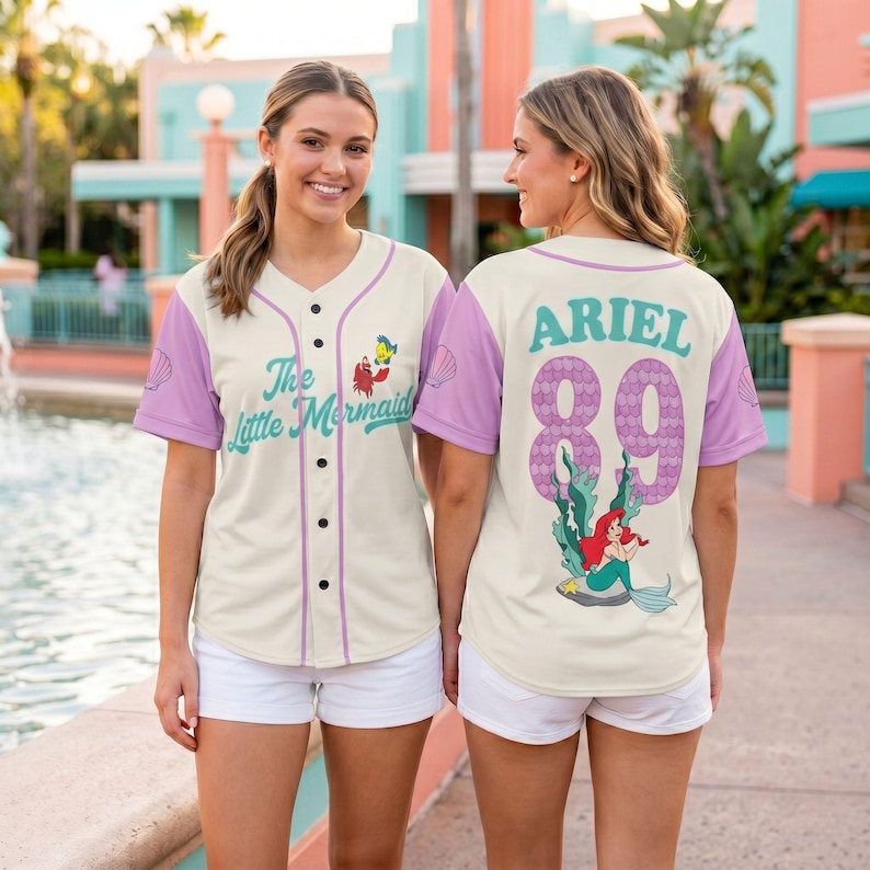 May include: Cream-colored baseball jersey with purple sleeves and accents. The front features the text "The Little Mermaid" and character graphics. The back displays "ARIEL 89" with a mermaid graphic. A fun, themed apparel item.