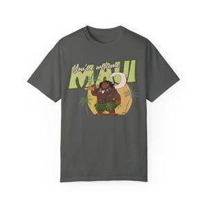 Puede incluir: Una camiseta gris oscuro con una imagen de dibujos animados de Maui de la pel&iacute;cula Moana. El texto "You're welcome Maui" est&aacute; impreso en la camiseta en un estilo retro.