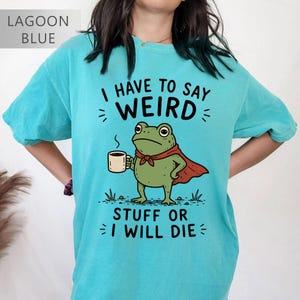 Puede incluir: Una camiseta azul laguna con un gráfico de una rana verde que lleva una capa roja y sostiene una taza de café. El texto dice I HAVE TO SAY WEIRD STUFF OR I WILL DIE. El diseño incluye pequeñas marcas negras y detalles de hierba.