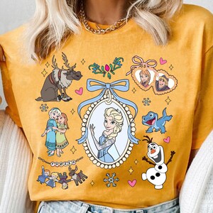 Puede incluir: Camiseta de color amarillo mostaza con ilustraciones de dibujos animados de personajes de la pel&iacute;cula animada "Frozen". El dise&ntilde;o incluye a Elsa, Anna, Olaf, Sven y Kristoff, junto con copos de nieve y corazones. Tambi&eacute;n est&aacute; presente la palabra "Frozen".