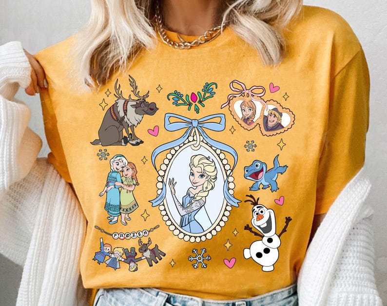 Puede incluir: Camiseta de color amarillo mostaza con ilustraciones de dibujos animados de personajes de la pel&iacute;cula animada "Frozen". El dise&ntilde;o incluye a Elsa, Anna, Olaf, Sven y Kristoff, junto con copos de nieve y corazones. Tambi&eacute;n est&aacute; presente la palabra "Frozen".