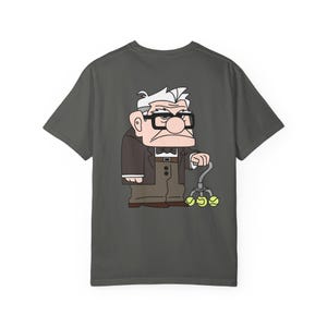May include: Dark gray t-shirt featuring a cartoon illustration of an elderly man with glasses, a bow tie, and a cane with tennis balls. The character is wearing a brown suit jacket and pants. The design is centered on the shirt.