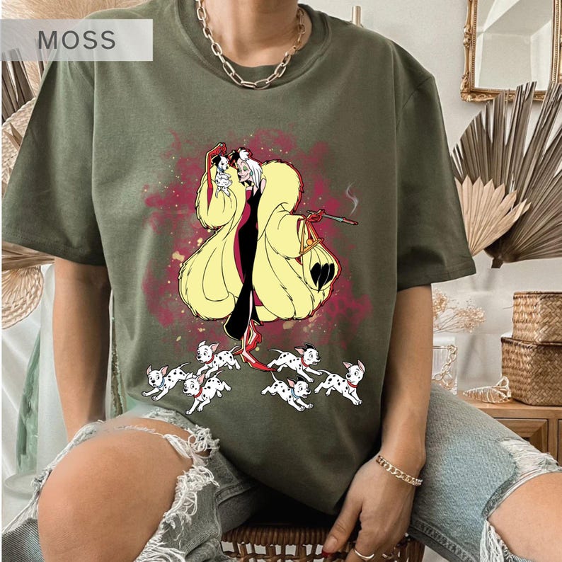 K&ouml;nnte beinhalten: Olivgr&uuml;nes T-Shirt mit einer Cartoon-Illustration einer Figur in einem gelben Mantel, einem schwarzen Kleid und roten Schuhen, die eine Zigarette und eine Handtasche h&auml;lt, mit Dalmatiner-Welpen. Das Wort "MOSS" befindet sich in der oberen linken Ecke.