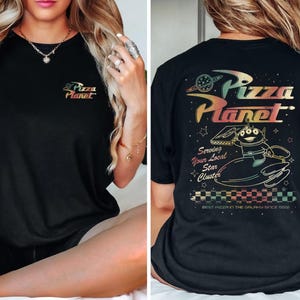 Puede incluir: Camiseta negra con un gr&aacute;fico colorido del logotipo de Pizza Planet y un personaje de la pel&iacute;cula Toy Story. El texto en la camiseta dice "Pizza Planet", "Serving Your Local Star Cluster", y "Best Pizza in the Galaxy Since 1995".