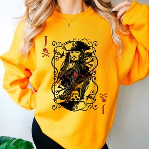 May include: Mustard yellow sweatshirt featuring a graphic of a playing card with a pirate design. The card shows a portrait of a pirate with long hair and a hat, surrounded by decorative elements, skulls, and the letter "J".