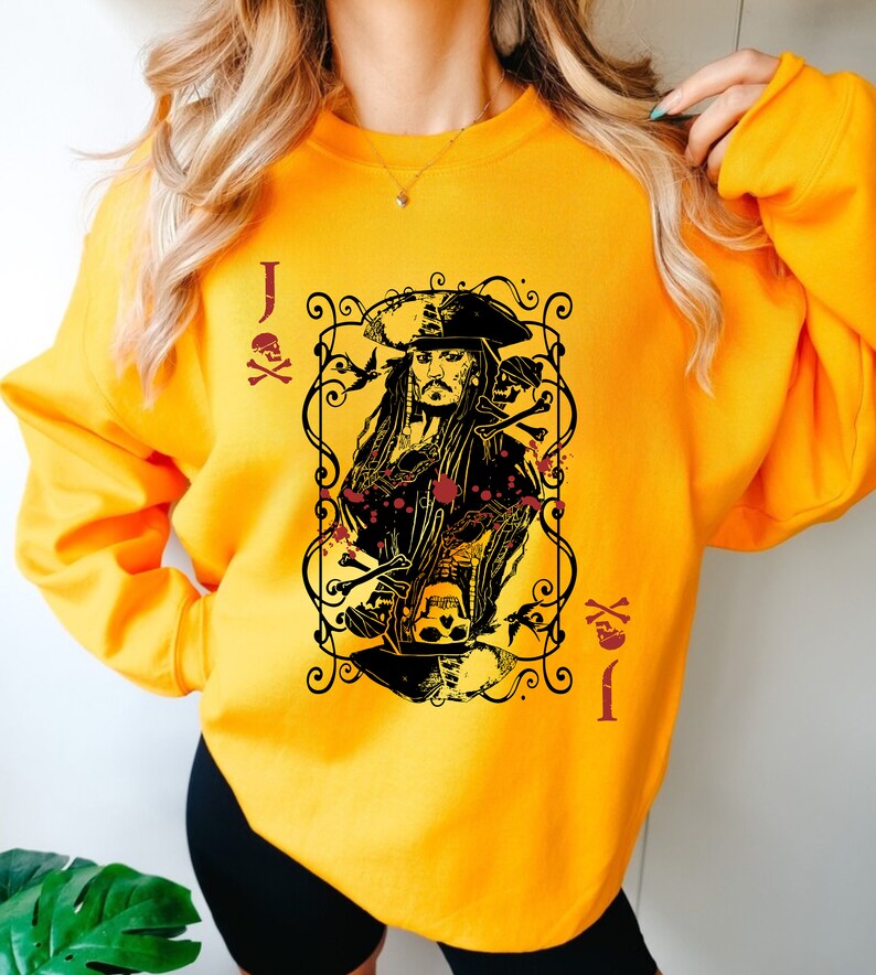 May include: Mustard yellow sweatshirt featuring a graphic of a playing card with a pirate design. The card shows a portrait of a pirate with long hair and a hat, surrounded by decorative elements, skulls, and the letter "J".