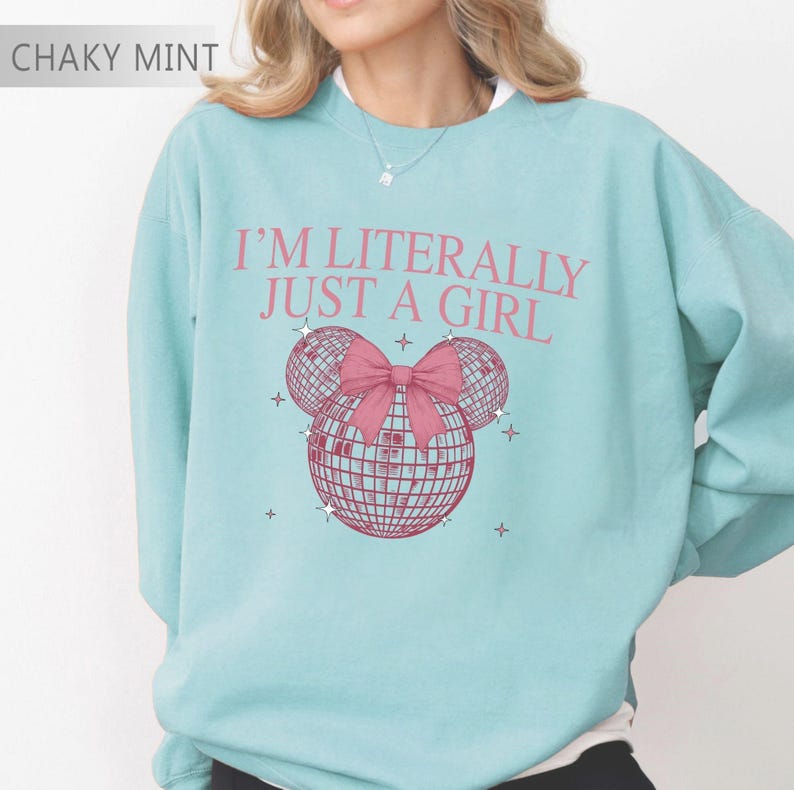 May include: A light blue sweatshirt with the text "I'M LITERALLY JUST A GIRL" in pink. The design features three disco ball shapes with a pink bow, and small star accents. The top left corner has the text "CHAKY MINT".