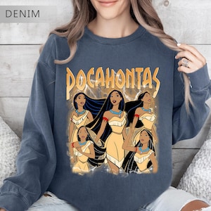 Pode incluir: Camisola azul denim com um gr&aacute;fico de Pocahontas e a palavra "Pocahontas" em letras douradas. O gr&aacute;fico mostra v&aacute;rias imagens de Pocahontas em um vestido amarelo com cabelo preto e um colar turquesa. A camisola tem gola redonda.