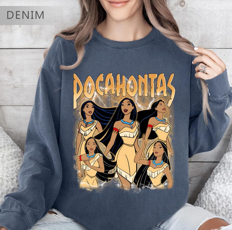 Pode incluir: Camisola azul denim com um gr&aacute;fico de Pocahontas e a palavra "Pocahontas" em letras douradas. O gr&aacute;fico mostra v&aacute;rias imagens de Pocahontas em um vestido amarelo com cabelo preto e um colar turquesa. A camisola tem gola redonda.