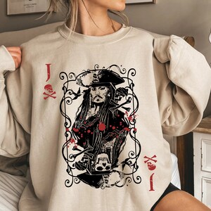 May include: Beige sweatshirt featuring a graphic of a pirate captain in a playing card design. The design includes skulls, crossbones, and the letter "J" with a skull and crossbones symbol in red. The sweatshirt has long sleeves.