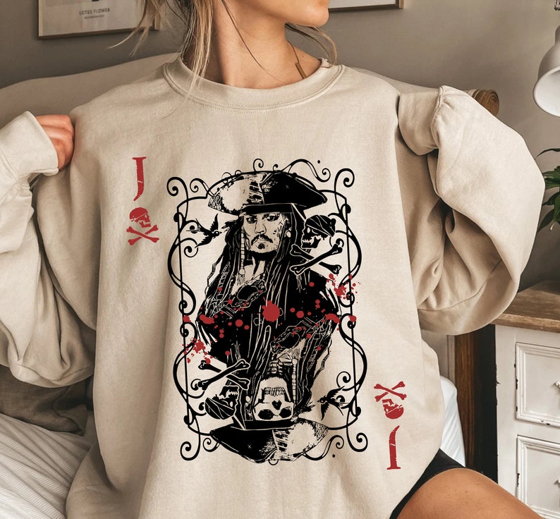 May include: Beige sweatshirt featuring a graphic of a pirate captain in a playing card design. The design includes skulls, crossbones, and the letter "J" with a skull and crossbones symbol in red. The sweatshirt has long sleeves.