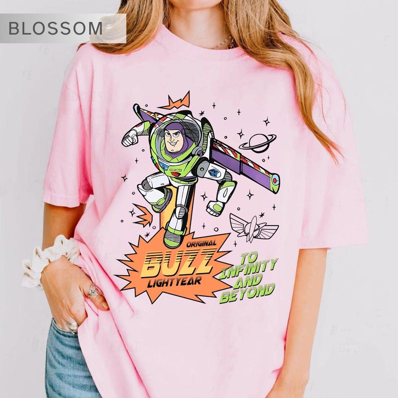 K&ouml;nnte beinhalten: Hellrosa T-Shirt mit einem Buzz Lightyear-Motiv. Das Design enth&auml;lt den Text "Original Buzz Lightyear" und "To Infinity and Beyond". Das T-Shirt hat einen Rundhalsausschnitt und ist im l&auml;ssigen Stil gehalten. Die Grafik ist in einem kr&auml;ftigen, farbenfrohen Stil gehalten.