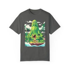 Puede incluir: Una camiseta gris oscuro con un estampado gr&aacute;fico colorido de una figura de diosa verde con una joven de pie frente a ella. La diosa est&aacute; rodeada de exuberante vegetaci&oacute;n verde y la mujer sostiene una lanza. El gr&aacute;fico est&aacute; sobre un fondo de agua azul y nubes blancas.