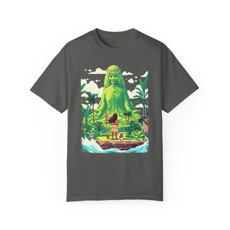 Puede incluir: Una camiseta gris oscuro con un estampado gr&aacute;fico colorido de una figura de diosa verde con una joven de pie frente a ella. La diosa est&aacute; rodeada de exuberante vegetaci&oacute;n verde y la mujer sostiene una lanza. El gr&aacute;fico est&aacute; sobre un fondo de agua azul y nubes blancas.