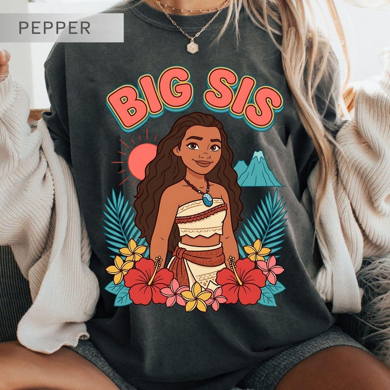 May include: Dark gray sweatshirt with a colorful graphic featuring the text "BIG SIS" above a cartoon illustration of a woman surrounded by flowers, palm leaves, a sun, and a mountain.