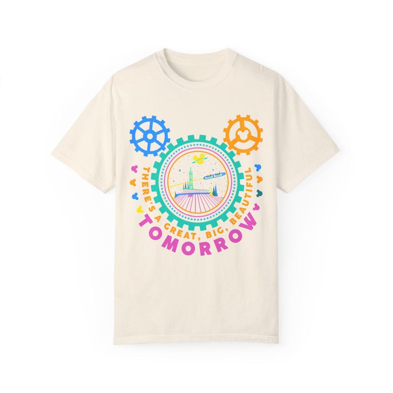 May include: A cream-coloured short-sleeve t-shirt featuring a graphic of a futuristic building inside a gear-shaped frame. The text "THERE'S A GREAT, BIG, BEAUTIFUL TOMORROW" is printed in pink, orange, and yellow around the central design.
