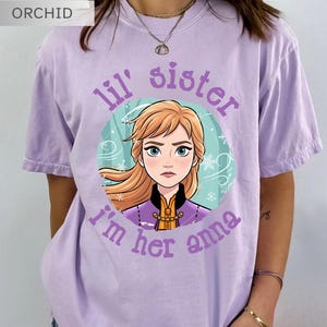 May include: Orchid purple t-shirt with a graphic of a cartoon character with blonde hair and blue eyes, surrounded by a teal circle with snowflakes. Text reads "lil' sister I'm her anna".