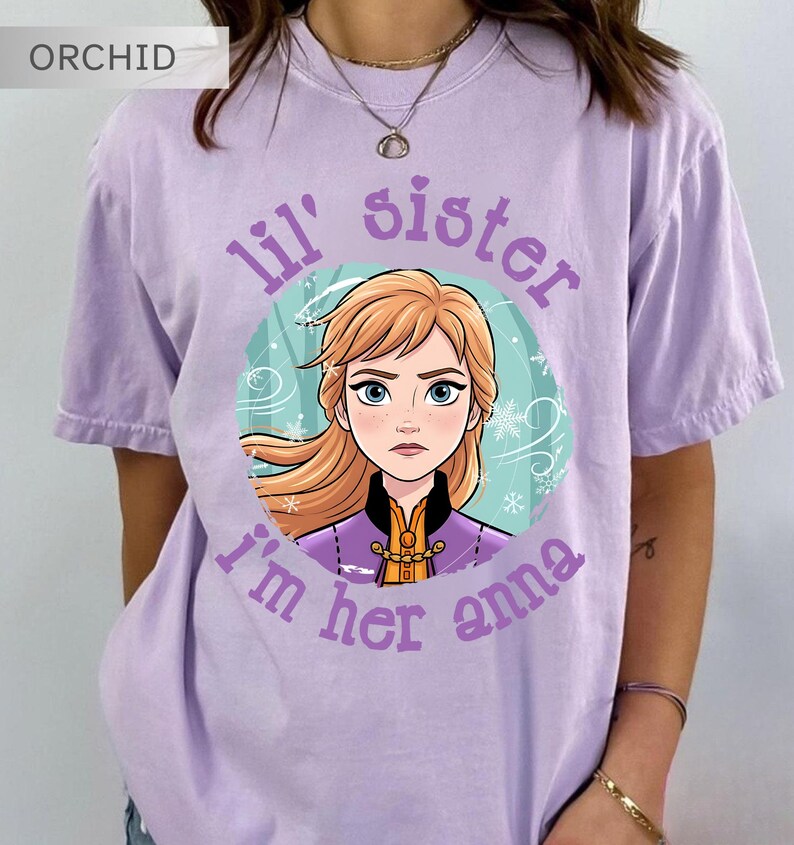 May include: Orchid purple t-shirt with a graphic of a cartoon character with blonde hair and blue eyes, surrounded by a teal circle with snowflakes. Text reads "lil' sister I'm her anna".