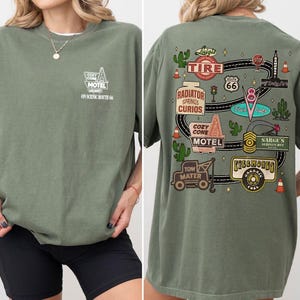 May include: An olive green t-shirt featuring a small white graphic on the front chest that reads Cozy Cone Motel On Scenic Route 66. The back displays a large, colorful illustration of a winding road with various signs and landmarks from Route 66.