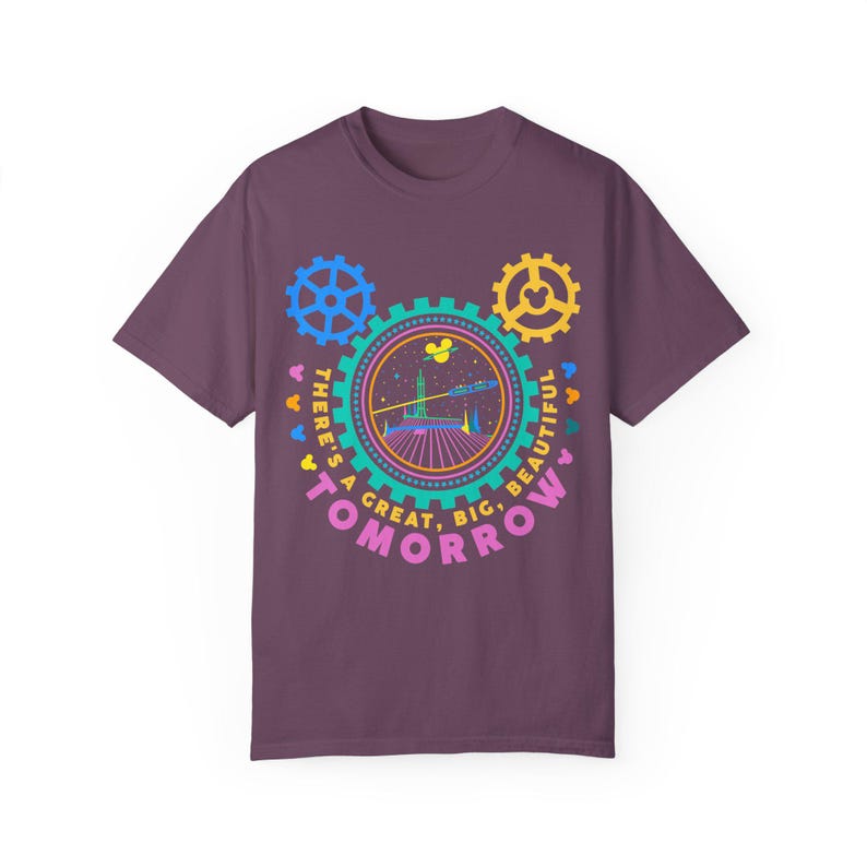 May include: A dark purple short-sleeve t-shirt featuring a graphic print of a central gear containing a space-themed illustration, flanked by two smaller gears. The text reads, "THERE'S A GREAT, BIG, BEAUTIFUL TOMORROW" in a circular arrangement.