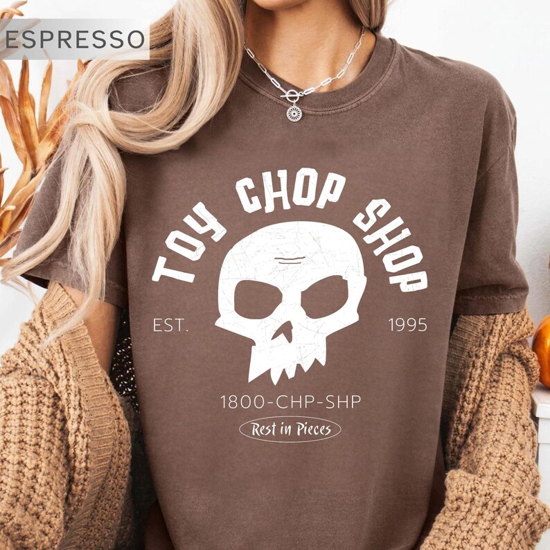 Pu&ograve; includere: Una t-shirt marrone a maniche corte con una grafica bianca raffigurante un teschio e il testo TOY CHOP SHOP, EST. 1995, 1800-CHP-SHP e Rest in Pieces. La maglia presenta un girocollo e un aspetto vissuto di ispirazione vintage.