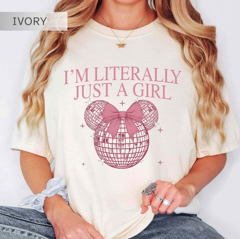 May include: Ivory-colored t-shirt with the text "I'M LITERALLY JUST A GIRL" in pink. The design features a pink bow and three disco ball shapes. The shirt is a casual, comfortable style.