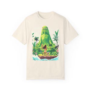Puede incluir: Una camiseta de color crema con una figura de diosa verde y una joven de pie frente a ella. La diosa est&aacute; rodeada de exuberante vegetaci&oacute;n verde y flores. La camiseta tiene un estilo de dibujos animados.