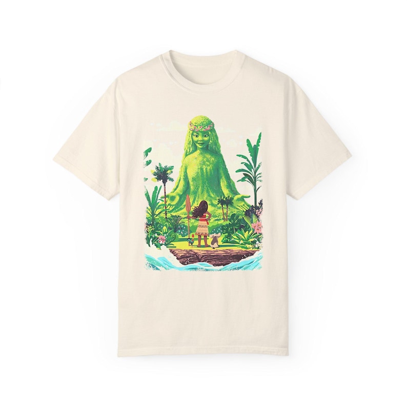 Puede incluir: Una camiseta de color crema con una figura de diosa verde y una joven de pie frente a ella. La diosa est&aacute; rodeada de exuberante vegetaci&oacute;n verde y flores. La camiseta tiene un estilo de dibujos animados.