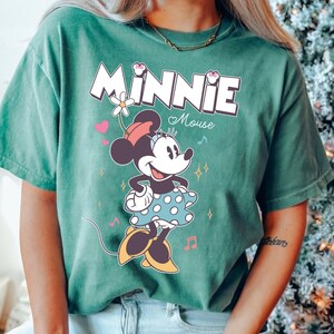 Puede incluir: Una camiseta verde con un gr&aacute;fico de Minnie Mouse con un vestido de lunares. El texto "Minnie Mouse" est&aacute; impreso en blanco sobre el gr&aacute;fico.