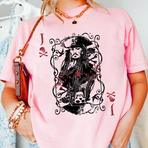 May include: A light pink t-shirt featuring a black and red graphic of a pirate card. The graphic includes a portrait of a pirate, skulls, and the letter "J". The shirt has a crew neck and short sleeves.