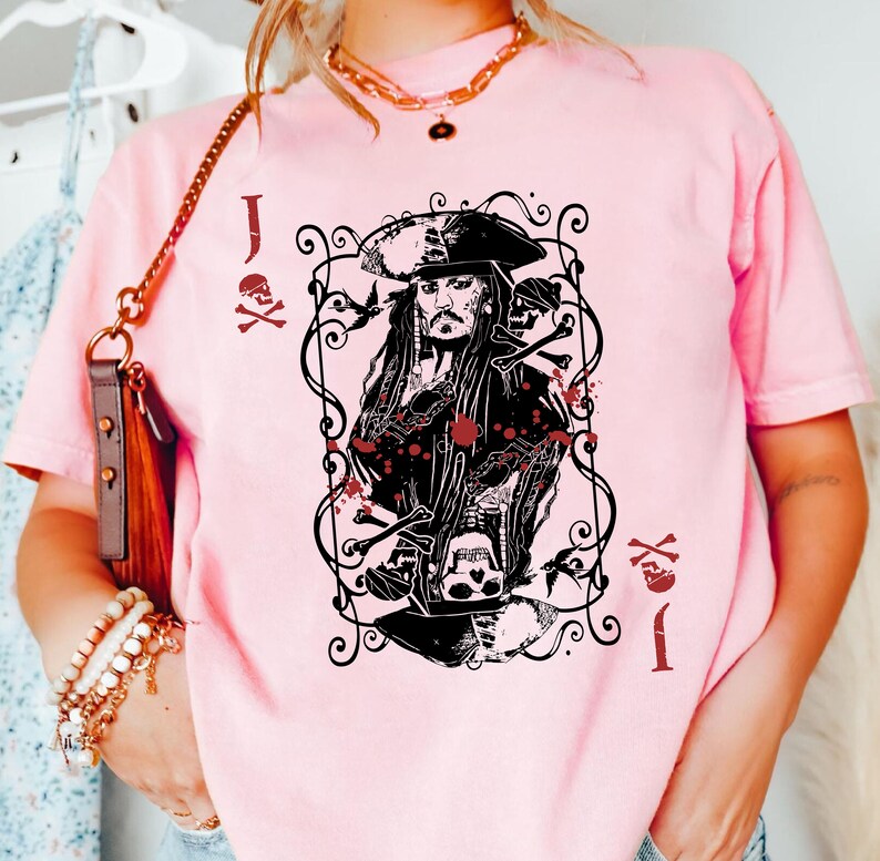 May include: A light pink t-shirt featuring a black and red graphic of a pirate card. The graphic includes a portrait of a pirate, skulls, and the letter "J". The shirt has a crew neck and short sleeves.
