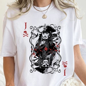 May include: White t-shirt featuring a black and red graphic of a playing card with a pirate design. The card shows a pirate with long hair and a hat, surrounded by decorative elements and the letter "J".