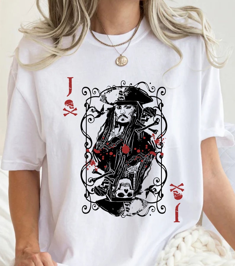 May include: White t-shirt featuring a black and red graphic of a playing card with a pirate design. The card shows a pirate with long hair and a hat, surrounded by decorative elements and the letter "J".