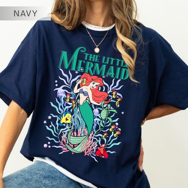 May include: Navy blue t-shirt featuring a graphic of Ariel from The Little Mermaid. The design includes Ariel with red hair and a green tail, surrounded by sea creatures and the text "The Little Mermaid" in green.
