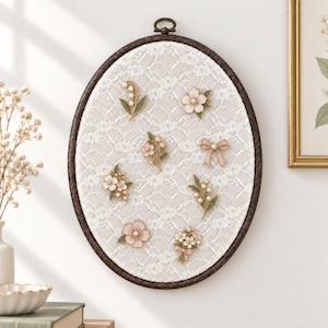 May include: An oval embroidery hoop display board covered in white lace fabric, featuring eight assorted floral and bow-shaped enamel pins. The pins include designs of lily of the valley, cherry blossoms, and a small pink ribbon bow, all arranged.