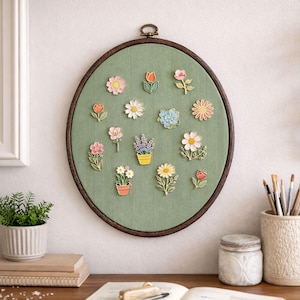 May include: An oval wall hanging with a green fabric backdrop and a dark brown wooden frame. The fabric displays various colourful flower pins, including daisies, tulips, and lavender. A decorative piece for a home or office.