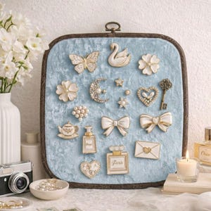 May include: A collection of decorative embellishments on a square, light blue velvet background. The embellishments include a butterfly, swan, flowers, bows, a key, and a perfume bottle with the word "Paris". The frame is a dark brown color.
