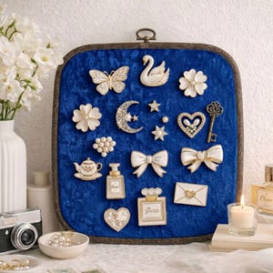 May include: A display of decorative brooches and pins on a blue velvet backdrop. The collection features a butterfly, swan, flowers, bows, a key, and perfume bottles. The background is framed in dark wood. A vintage camera and a vase of white flowers are in the background.