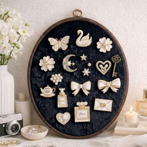 May include: An oval wooden embroidery hoop display board featuring a black velvet backing. Attached to the velvet are various gold-toned and white enamel pins, including a swan, butterfly, crescent moon, heart, bow, teapot, perfume bottles, and envelope.