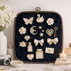 May include: A collection of decorative pins and brooches arranged on a dark blue velvet display. The pins feature various designs, including a butterfly, swan, flowers, bows, a crescent moon, stars, a key, a teapot, perfume bottles, and hearts. The pins are white and gold.