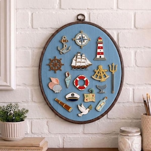 Könnte beinhalten: Wandbehang mit nautischem Thema mit Metallanhängern auf blauem Stoff. Zu den Anhängern gehören ein Anker, ein Kompass, ein Leuchtturm, ein Steuerrad, ein Segelboot, eine Rettungsboje und Muscheln. Der Rahmen ist oval und dunkelbraun.
